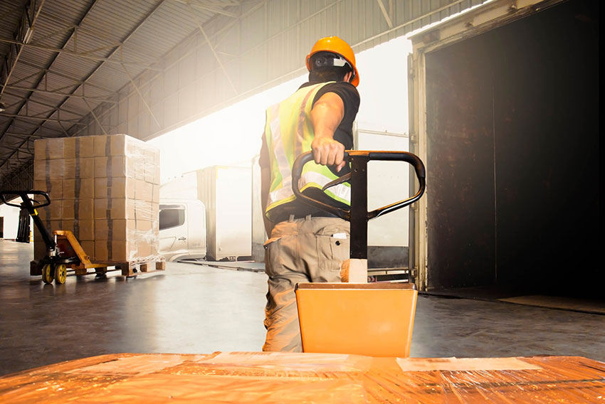 A man with a hardhat pulling a dolly in a warehouse
